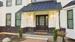 Modern house entrance with black double doors, white siding, black metal roof accents, brick and stone trim, two wall lanterns, potted plants, and a doormat on a tiled porch. House number 102 is visible by the door.